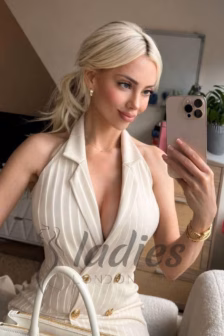 Blonde woman in a cream pinstripe blazer dress takes a mirror selfie, holding a gold iPhone.
