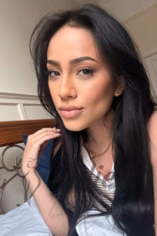 Close-up portrait of a woman with long black hair, wearing a white top and gold necklace, resting on a bed in a softly lit room.