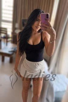 Woman taking a mirror selfie in a living room, wearing a black tank top and beige mini skirt.