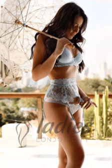 Woman in light blue lace lingerie standing on a balcony, holding a lace parasol with a cityscape in the background.