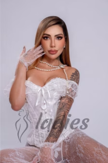 Portrait of a tattooed woman in a white lace corset with a pearl necklace and lace gloves, posed against a gray backdrop.