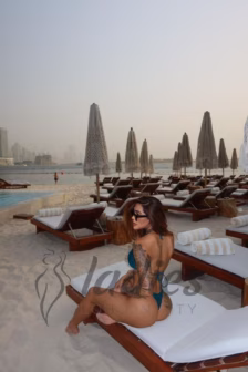 Woman in a teal bikini sits on a white pool lounge on a sandy beach, with tattooed arms and sunglasses, rows of closed umbrellas behind her.