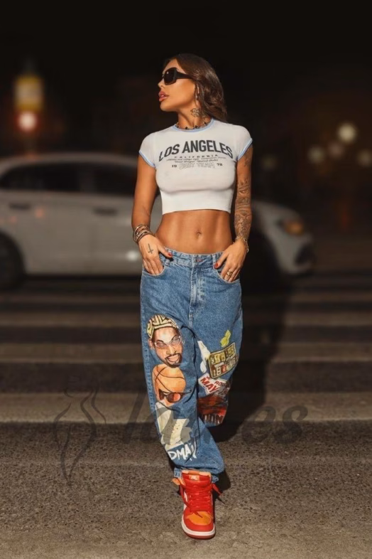Fashionable woman posing on a nighttime street wearing a white crop top that says 'LOS ANGELES', oversized jeans with cartoon graffiti, red high-top sneakers, and dark sunglasses; tattoos visible on her arms.