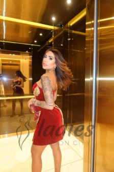 Woman in a red dress with gold embellishments posing in a gold-toned mirrored elevator lobby; tattoos visible on her left arm.