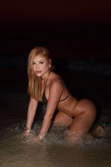 Woman in a black bikini kneeling in shallow water at night, facing the camera with blonde hair.