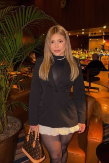 Woman in a black blazer with a sheer top and white ruffled skirt posing in a bar/lounge, holding a brown LV handbag.
