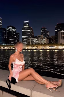 Woman in a pink satin dress sits on a concrete ledge by a nighttime waterfront with a city skyline behind her.