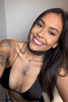 Smiling woman taking a selfie in a black bikini top, with visible arm and chest tattoos and a studded choker necklace.