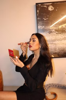 Woman in a black dress sits at a vanity applying lipstick while holding a red compact mirror open to her lips.