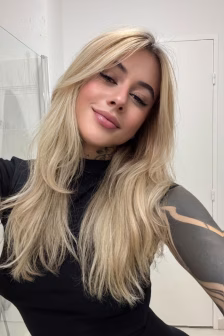 Self-portrait of a woman with long blonde hair and a small smile, wearing a black top in a bright bathroom, showing a neck tattoo.