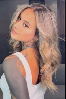 Woman with wavy blonde hair and visible neck tattoo, wearing a white tank top, looking over her shoulder at the camera.