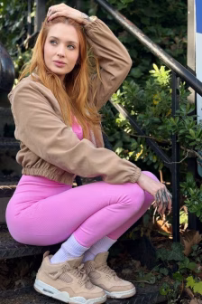 Woman with long red hair squats on outdoor stairs, wearing a tan bomber jacket, pink leggings, and beige sneakers, looking to the side.