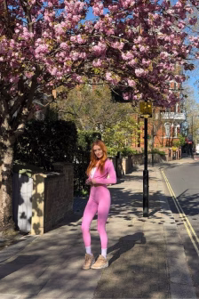 Person wearing a bright pink one-piece outfit stands on a sidewalk beneath a flowering pink tree on a sunny street.