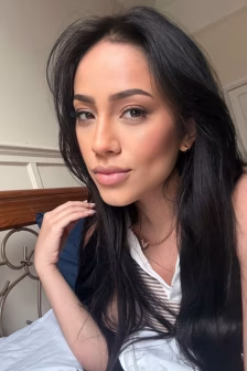 Close-up portrait of a woman with long black hair, wearing a white top and gold necklace, resting on a bed in a softly lit room.
