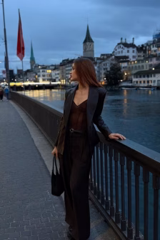 Woman in a dark suit and brown top stands by a railing along a river at dusk, holding a black handbag, with a European cityscape in the background.