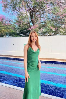 Woman in a silky green gown posing beside a blue-tiled pool with a flowering tree in bloom behind her, sunny day.