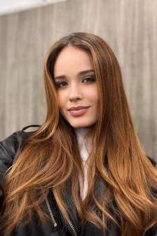 Young woman with long, light brown hair wearing a black leather jacket and a light-colored top, looking at the camera with a slight smile.