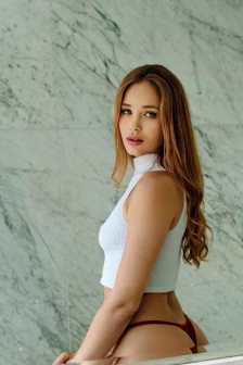 Portrait of a woman with long red hair wearing a white ribbed sleeveless crop top and burgundy thong, against a marble wall.