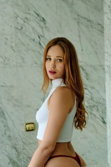 Portrait of a red-haired woman in a white ribbed sleeveless top and burgundy bikini bottoms, posing sideways against a marble wall.