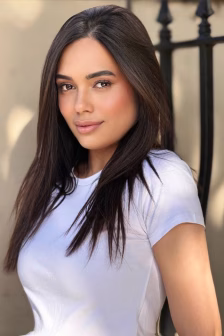 Portrait of a woman with long dark hair wearing a white T‑shirt, standing beside a black iron fence outdoors.