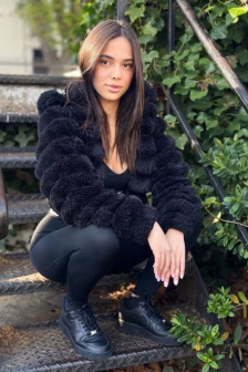 Woman crouching on outdoor metal stairs in a black fur coat, black leggings, and sneakers, hands resting on knees amid greenery