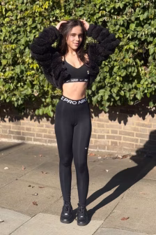 Woman in a black Nike athletic outfit poses outdoors, hands on head, in front of a leafy hedge.