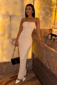 Woman in a white embellished strapless dress stands beside a marble counter in a warmly lit luxe room, holding a black handbag with a chain strap.