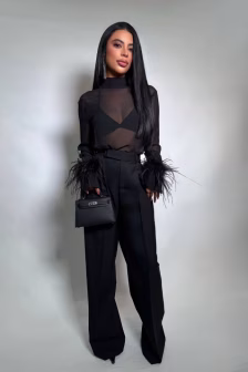 Woman in a black sheer top with feathered cuffs, wide-leg black trousers, and a small black handbag, standing against a neutral backdrop.