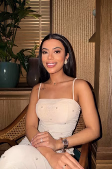 Woman with long dark hair in a white beaded spaghetti-strap dress, sitting on a wooden chair and smiling.