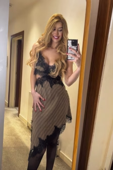 Woman in a beige and black lace slip dress posing in a mirror selfie with a smartphone held in her right hand