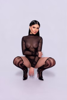 Woman in a sheer black bodysuit and striped stockings crouches forward with hands between her knees in a studio backdrop.