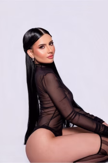Woman with long black hair in a sheer black bodysuit seated sideways, looking toward the camera against a light background.