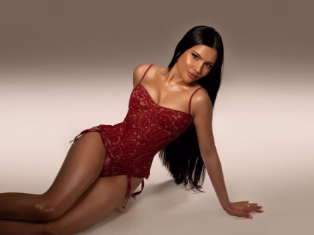 Model wearing red lace lingerie reclining on a beige studio backdrop, looking at the camera with long dark hair flowing down. Alternative: Woman in burgundy corset posing on the floor for a lingerie shoot.