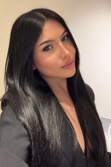 Portrait of a woman with long, glossy black hair wearing a black blazer, posing against a light wall in a selfie-style photo.