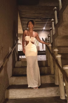 Smiling woman wearing a cream crochet two-piece dress stands on a wooden staircase in a rustic interior.