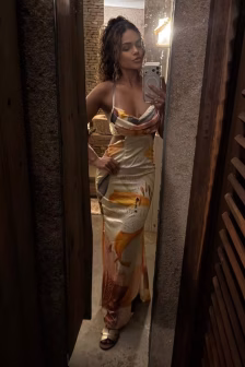 Woman posing in a colorful strapless maxi dress, taking a mirror selfie with her phone in a rustic, narrow doorway.