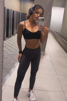 Woman in a black sports bra and leggings taking a mirror selfie in a gym, wearing headphones and white sneakers.