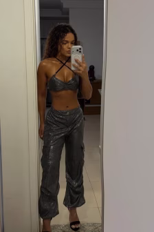 Full-length mirror selfie of a person wearing a metallic gray sparkly two-piece outfit (crisscross halter top and high-waisted cropped pants) standing in a doorway, holding a phone.