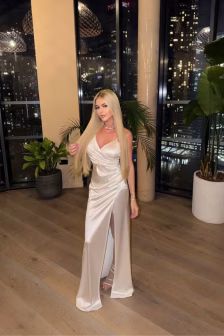 Woman with long blonde hair in a white satin gown with a thigh-high slit, posing indoors near large windows with city lights visible outside.