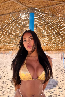 Woman in a yellow bikini posing on a sandy beach under a woven straw umbrella with a blue pole, facing the camera.