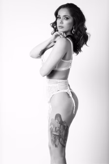 Portrait of a woman in white lingerie posing in profile, a large thigh tattoo visible on her leg.