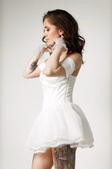 Woman in a white off-shoulder wedding dress, adjusting dotted sheer gloves, tattoos visible on her thigh as she faces left in profile.