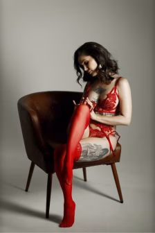 Woman in red lace lingerie and thigh-high stockings sits on a brown chair, looking down at her legs with tattoos visible on her torso and thigh.