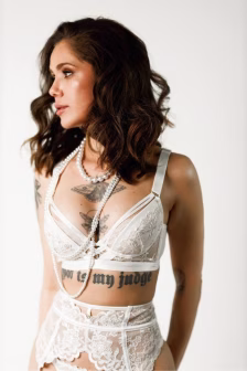 Woman in white lace lingerie wearing layered pearl necklaces, tattoos on chest and abdomen visible as she looks to the left.