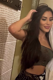 Woman with long dark hair in an olive green crop top with cutouts, posing with her hand on her head in a tiled bathroom.