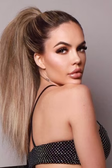Glamour portrait of a woman with a high blonde ponytail, bold makeup, wearing a black polka-dot top photographed from the side.