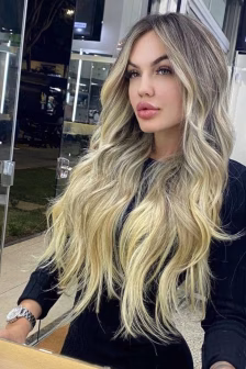 Woman with long wavy blonde hair wearing a black top sits in a salon, looking at the camera.