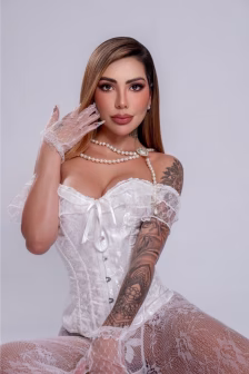 Portrait of a tattooed woman in a white lace corset with a pearl necklace and lace gloves, posed against a gray backdrop.