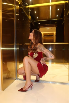 Woman in a red velvet dress with gold embellishments crouches in a gold-toned elevator lobby, showcasing tattoos on her left arm.