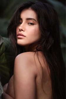Close-up portrait of a woman with long dark hair and bare shoulders, gazing softly at the camera with a green, leafy background behind her.